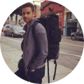 Man with a large backpack walking on a city street with cars and buildings in the background.